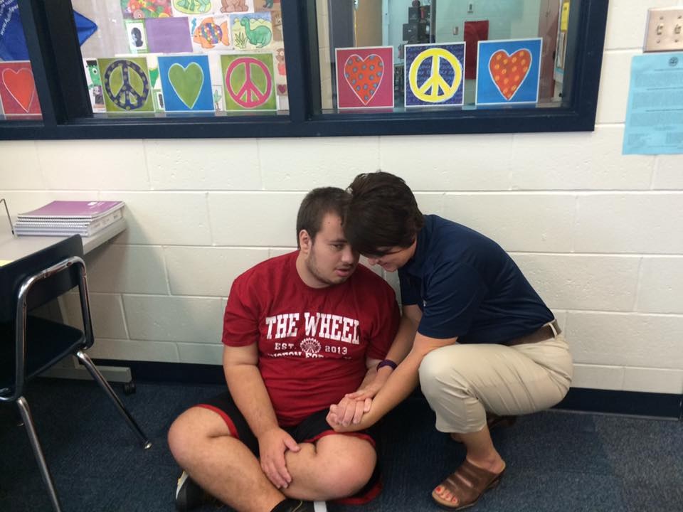 Photo of a young adult boy sitting cross leg on a classroom floor, with an aid talking kindly to him